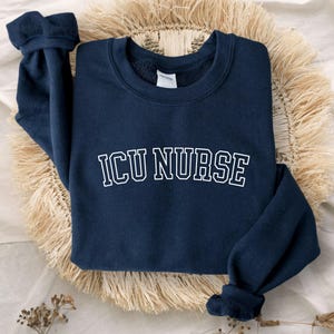 May include: A navy blue long-sleeve sweatshirt featuring the text ICU NURSE in a white, arched, collegiate-style font across the chest. The garment has a crew neckline and ribbed cuffs, providing a simple and functional design for medical professionals.