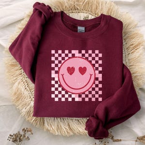 May include: A maroon long-sleeve sweatshirt featuring a central graphic of a pink smiley face with heart-shaped eyes. The smiley face is set against a white and pink checkered square background. The design has a distressed, weathered texture.