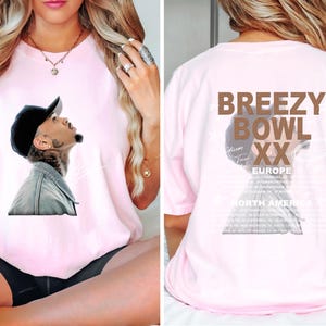 May include: A light pink t-shirt featuring a graphic of a man looking upwards, wearing a black cap and denim jacket. The back of the shirt displays the text "BREEZY BOWL XX EUROPE" and a list of tour dates.