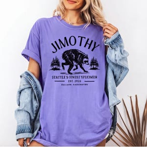 A woman wearing a purple t-shirt with a graphic design of a bear and text that says "Jimthy" and "SEATTLE'S FINEST SPECIMEN EST. 1826 BALLARD, WASHINGTON".