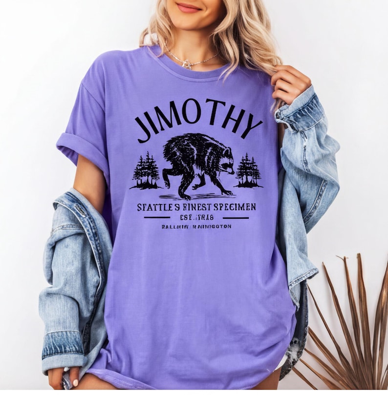 A woman wearing a purple t-shirt with a graphic design of a bear and text that says "Jimthy" and "SEATTLE'S FINEST SPECIMEN EST. 1826 BALLARD, WASHINGTON".
