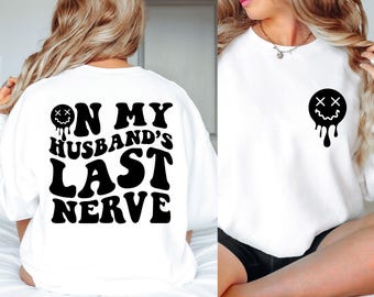 May include: A white sweatshirt featuring a black graphic on the front left chest of a melting smiley face with X eyes. The back displays the black text ON MY HUSBAND'S LAST NERVE in a bold, wavy font, accompanied by the same melting smiley face icon.