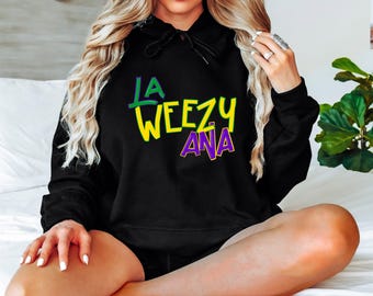 May include: A black hooded sweatshirt featuring the text LA WEEZY ANA printed on the front. The word LA is in green, WEEZY is in yellow, and ANA is in purple with a yellow outline. The hoodie includes a drawstring hood and long sleeves.