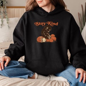 May include: Black hoodie with a front graphic of a brown monkey hugging a teddy bear. The text "Stay Kind" is arched above the graphic in orange. The hoodie has a kangaroo pocket and a drawstring hood.