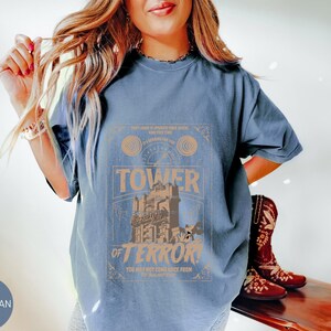 May include: Blue jean colored t-shirt with a graphic that reads "Tower of Terror". The graphic is in shades of tan and brown, featuring a tower and the words "You may not come back from". The shirt is a relaxed fit.