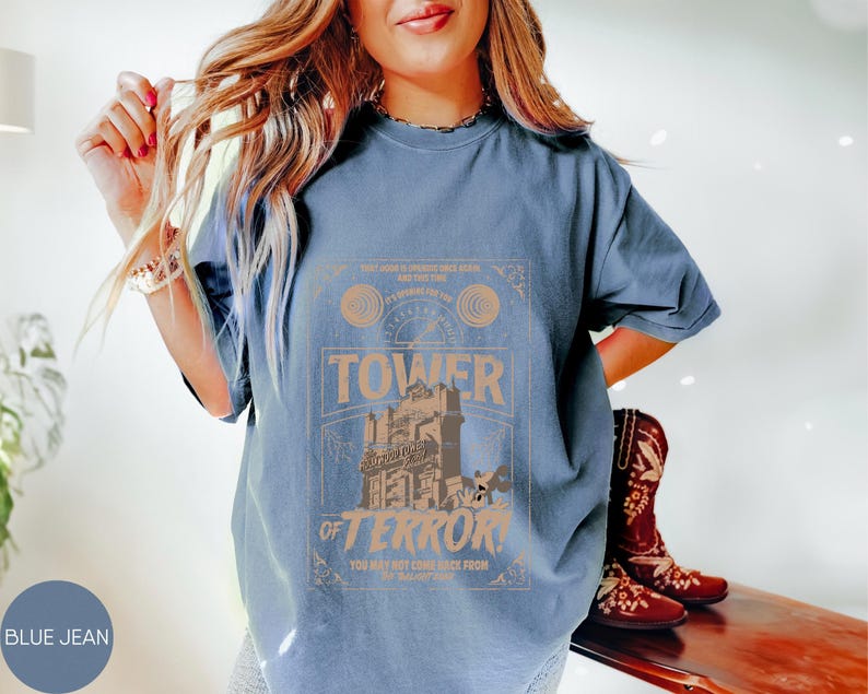 May include: Blue jean colored t-shirt with a graphic that reads "Tower of Terror". The graphic is in shades of tan and brown, featuring a tower and the words "You may not come back from". The shirt is a relaxed fit.