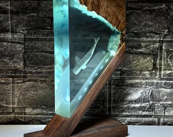 May include: A wooden table lamp with a clear resin base featuring a silhouette of a fish swimming in a blue and white ocean scene. The lamp has a natural wood finish and a triangular shape.