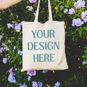 May include: A white canvas tote bag with the text "YOUR DESIGN HERE" printed in teal blue. The bag is held by a hand in front of a bush of purple flowers.