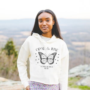 May include: Cream-colored cropped hoodie with the text "OPAL & JUNE" above a butterfly design and the words "YOUR DESIGN HERE". The hoodie is paired with a floral print skirt. The background is a blurred outdoor scene.