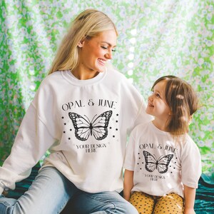 K&ouml;nnte beinhalten: Ein wei&szlig;es Sweatshirt und T-Shirt mit einem schwarzen Schmetterlingsmotiv und dem Text "Opal & June" und "Your Design Here".