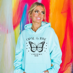 Puede incluir: Una sudadera con capucha azul claro con un gran diseño de mariposa en blanco y negro, el texto "OPAL & JUNE" y "YOUR DESIGN HERE". La sudadera tiene un bolsillo delantero y una capucha.