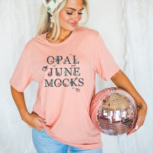 May include: A woman wearing a pink t-shirt with the text "OPAL and JUNE MOCKS" in black lettering with floral accents and butterflies. She is also wearing a white crocheted headband with pink and green flowers and blue jeans. She is holding a rose gold disco ball.