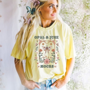 Pu&ograve; includere: Una maglietta gialla con un design grafico che presenta una farfalla, fiori e il testo "OPAL & JUNE MOCKS".