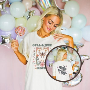 May include: A white t-shirt with a vintage-inspired graphic featuring a butterfly, flowers, and the text "Opal & June" and "Mochi". The back of the shirt has the text "Opal & June" in a cursive font.