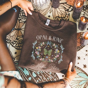 May include: A brown sweatshirt with the text "Opal & June" and a floral design with butterflies. The text "Mockup Photos" is also visible on the sweatshirt.