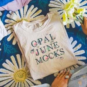 Puede incluir: Una camiseta beige con un diseño floral y el texto "OPAL and JUNE MOCKS" impreso en la parte delantera.