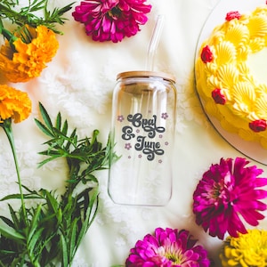 May include: A clear glass reusable water bottle with a bamboo lid and a straw. The bottle has the text "Cool & June" printed on it in black with pink flowers. The bottle is surrounded by yellow and pink flowers.
