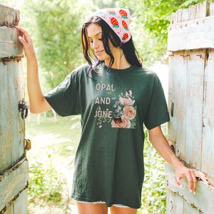 Pu&ograve; includere: Una maglietta verde scuro con un disegno floreale e il testo "Opal and June" stampato sul davanti. La modella indossa un cappello di uncinetto rosa con fragole.