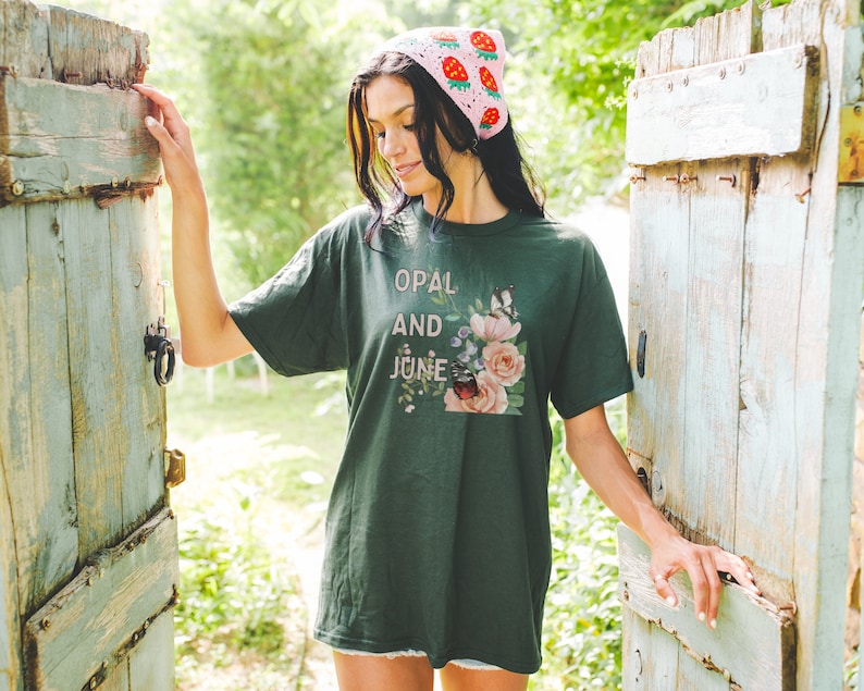 Pu&ograve; includere: Una maglietta verde scuro con un disegno floreale e il testo "Opal and June" stampato sul davanti. La modella indossa un cappello di uncinetto rosa con fragole.