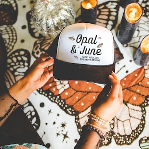 May include: White trucker hat with black mesh back. The hat has a black and white butterfly graphic and the text "Opal & June" with the tagline "Happy Mockups for Happy Designs".