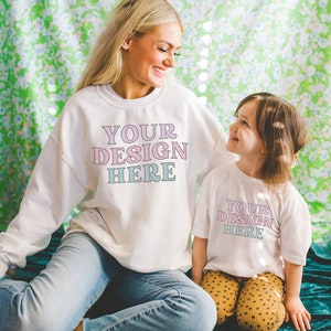 K&ouml;nnte beinhalten: Eine Frau und ein junges M&auml;dchen tragen wei&szlig;e Sweatshirts mit dem Text "YOUR DESIGN HERE" in rosa und blauen Buchstaben. Die Frau tr&auml;gt eine blaue Jeans und das M&auml;dchen tr&auml;gt eine gelbe Polka-Dot-Hose. Sie sitzen auf einer gr&uuml;nen, floral gemusterten Oberfl&auml;che.