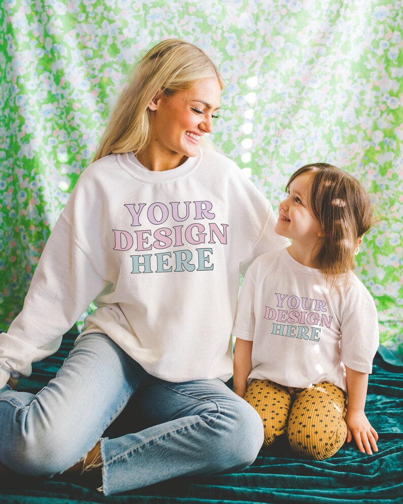 K&ouml;nnte beinhalten: Eine Frau und ein junges M&auml;dchen tragen wei&szlig;e Sweatshirts mit dem Text "YOUR DESIGN HERE" in rosa und blauen Buchstaben. Die Frau tr&auml;gt eine blaue Jeans und das M&auml;dchen tr&auml;gt eine gelbe Polka-Dot-Hose. Sie sitzen auf einer gr&uuml;nen, floral gemusterten Oberfl&auml;che.
