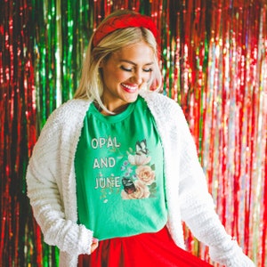 May include: A woman wearing a green t-shirt with the text "OPAL AND JUNE" and a floral design. She is also wearing a white fluffy cardigan and a red headband.