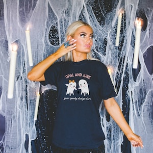Puede incluir: Una camiseta negra con el texto "Opal and June" y un gráfico de dos fantasmas que se toman de la mano con una araña entre ellos. El texto "your spooky design here" está debajo de los fantasmas.