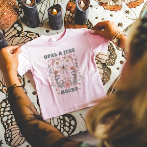 Mockup di maglietta rosa: foto colorata del modello che piega la maglietta in jersey fine per neonati Pink Rabbit Skins 3322 / Mockup di Whimsigoth accogliente per Halloween