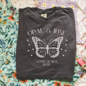 May include: A dark gray t-shirt with the text "OPAL & JUNE" and a white butterfly graphic. The text "YOUR DESIGN HERE" is below the butterfly.
