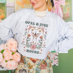 Puede incluir: Una sudadera gris con un diseño gráfico que presenta una mariposa, flores y hongos. El texto "OPAL & JUNE MOCKS" está impreso en la parte delantera de la sudadera.