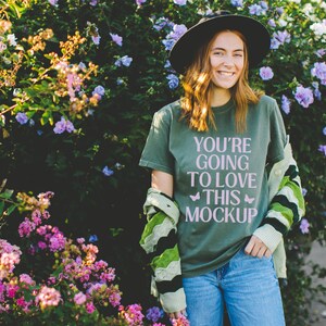 Op de afbeelding: Olijfgroen t-shirt met de roze tekst "YOU'RE GOING TO LOVE THIS MOCKUP". De persoon draagt een zwarte hoed, een blauwe jeans en een groen-wit patroon vest. De achtergrond toont kleurrijke bloemen.