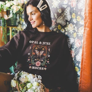 May include: A dark brown sweatshirt with the text "OPAL & JUNE MOCKS" and a floral and butterfly design. The person in the image is wearing pearl hair clips and holding a bouquet of flowers. The background features floral wallpaper.
