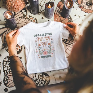 Mockup di camicia bianca: foto colorata del modello che piega la maglietta in jersey fine per neonati White Rabbit Skins 3322 / Mockup di Whimsigoth accogliente per Halloween