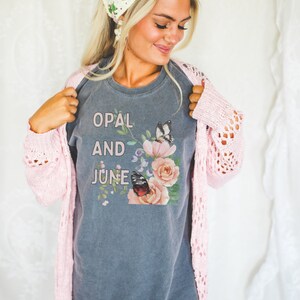 Puede incluir: Una camiseta gris con las palabras "OPAL AND JUNE" estampadas en la parte delantera, junto con un dise&ntilde;o floral y de mariposas. La persona lleva una rebeca rosa de punto abierto y un pa&ntilde;uelo de ganchillo blanco y rosa.