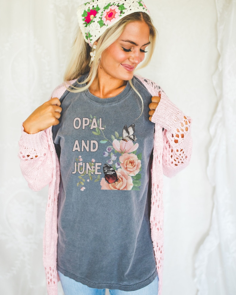 Puede incluir: Una camiseta gris con las palabras "OPAL AND JUNE" estampadas en la parte delantera, junto con un dise&ntilde;o floral y de mariposas. La persona lleva una rebeca rosa de punto abierto y un pa&ntilde;uelo de ganchillo blanco y rosa.