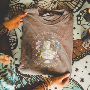 K&ouml;nnte beinhalten: Ein braunes T-Shirt mit einem grafischen Design eines B&uuml;stenbildes, das von Blumen umgeben ist. Der Text "OPAL & JUNE MOCKUPS" ist auf das T-Shirt gedruckt.