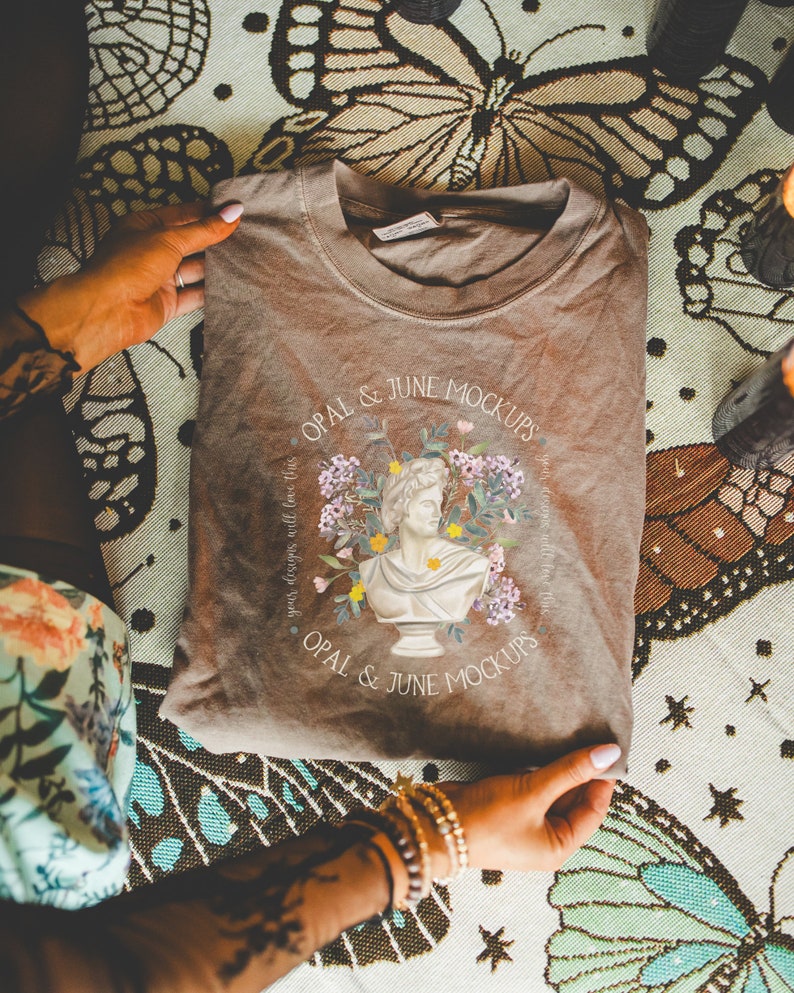 K&ouml;nnte beinhalten: Ein braunes T-Shirt mit einem grafischen Design eines B&uuml;stenbildes, das von Blumen umgeben ist. Der Text "OPAL & JUNE MOCKUPS" ist auf das T-Shirt gedruckt.