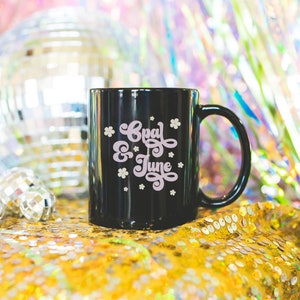 May include: Black ceramic mug with a white design that says "Opal & June" in a retro font. The mug is sitting on a gold sequined surface.