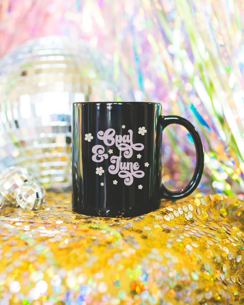 May include: Black ceramic mug with a white design that says "Opal & June" in a retro font. The mug is sitting on a gold sequined surface.
