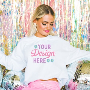 Pu&ograve; includere: Una felpa bianca con il testo "YOUR Design HERE" in lettere rosa e viola. La felpa &egrave; indossata da una persona davanti a uno sfondo argentato e iridescente.