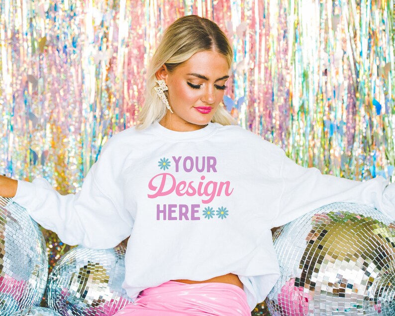 Pu&ograve; includere: Una felpa bianca con il testo "YOUR Design HERE" in lettere rosa e viola. La felpa &egrave; indossata da una persona davanti a uno sfondo argentato e iridescente.