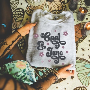 Może przedstawiać: Szara bluza z napisem "Opal & June" w retro czcionce z różowymi kwiatami wokół tekstu.