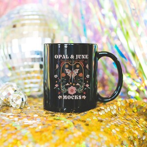 May include: Black ceramic mug with a white graphic design featuring a butterfly, flowers, and the text "OPAL & JUNE MOCKS". The mug is sitting on a gold sequined surface.