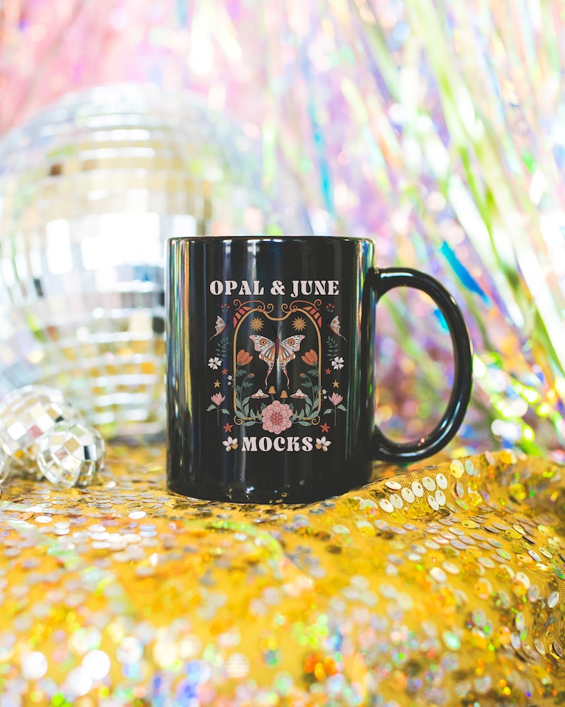 May include: Black ceramic mug with a white graphic design featuring a butterfly, flowers, and the text "OPAL & JUNE MOCKS". The mug is sitting on a gold sequined surface.