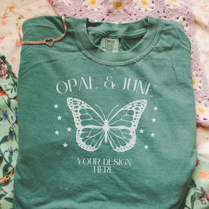 May include: A teal green t-shirt with the text "OPAL & JUNE" and a white butterfly graphic. The text "YOUR DESIGN HERE" is below the butterfly.