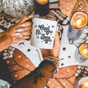 May include: White ceramic mug with a black and white graphic that says "Cozy & June" with pink flowers. The mug is being held by a person's hand.
