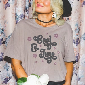 Puede incluir: Una camiseta marrón claro con el texto "Opal & June" en una fuente retro. El texto está rodeado de pequeñas flores rosas.