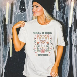 Pode incluir: Uma camiseta branca com um gr&aacute;fico de estilo vintage que apresenta uma borboleta, flores e o texto "OPAL & JUNE MOCKS".