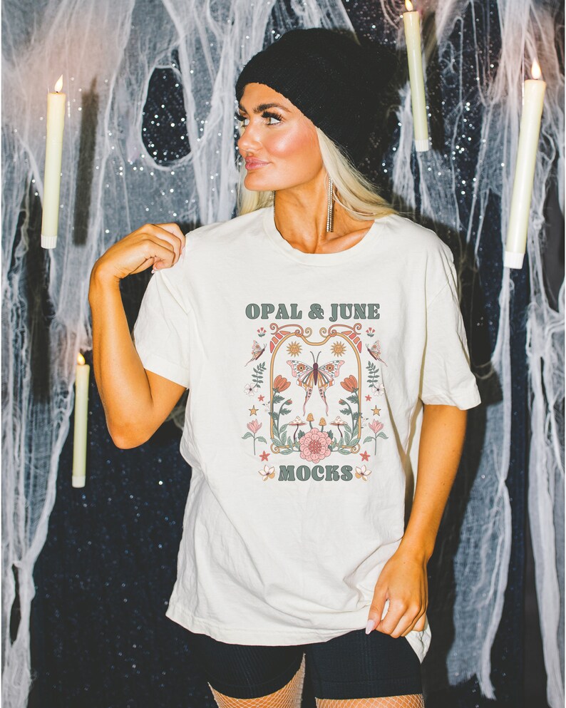 Pode incluir: Uma camiseta branca com um gr&aacute;fico de estilo vintage que apresenta uma borboleta, flores e o texto "OPAL & JUNE MOCKS".